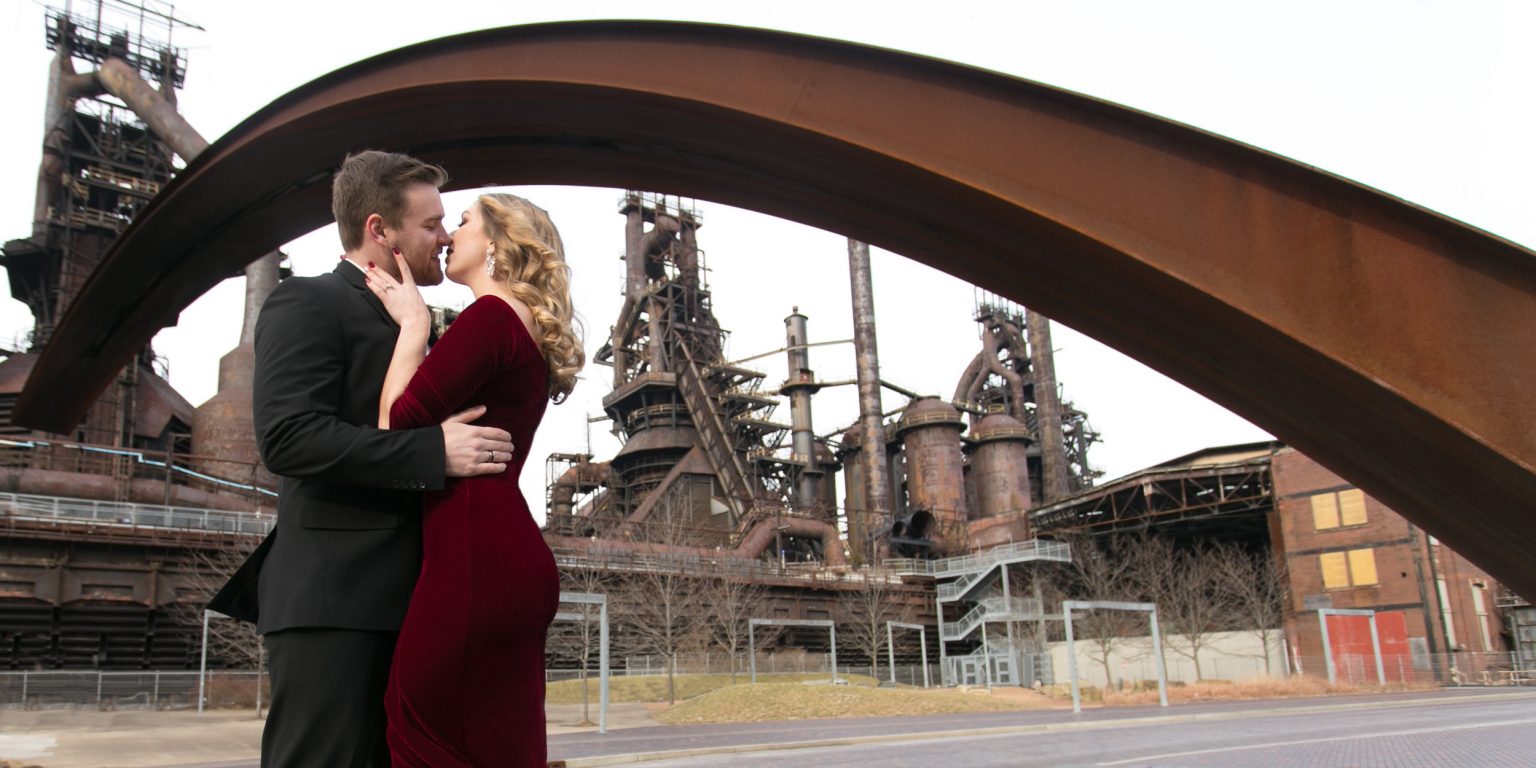 Fancy Dressed Engaged Couple kissing under artistic metal arch at Steel Stacks