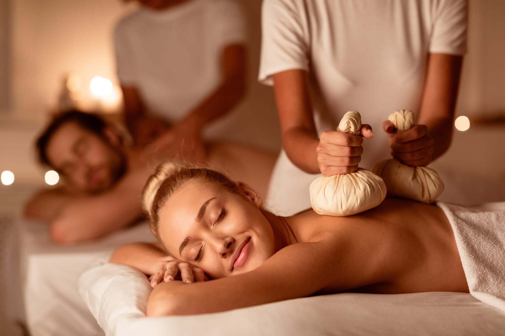 Relaxed engaged couple At Exotic Spa Resort, Lying With Eyes Closed Receiving Thai Massage With Aromatic Bags. Body Beauty Treatment, Relaxation And Wellness