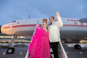 Couple in 1960's garb standing in TWA Hotel tarmac in front of a "Connie" TWA propeller airplane