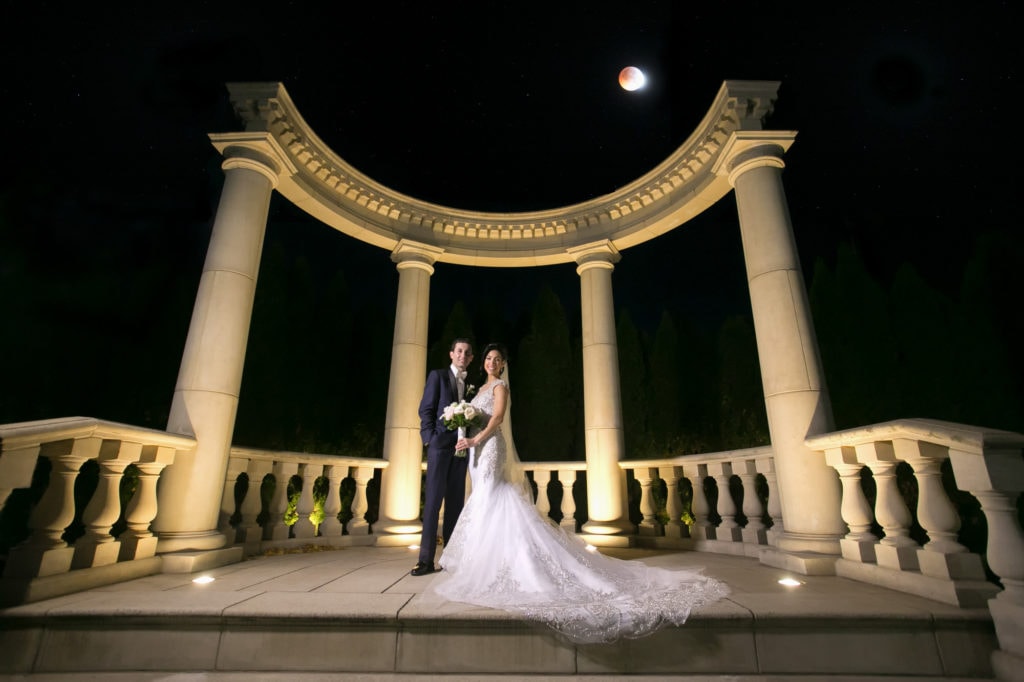 Bride and groom standing outdoors under greek /roman columns under and red moon at the the rockleigh